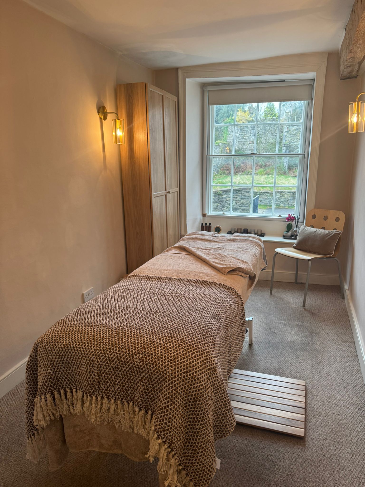 Massage couch dressed with a knitted throw, a wooden step and a chair by the window